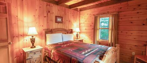A wooden cabin bedroom with a bed, two bedside tables with lamps, a window, and a door.