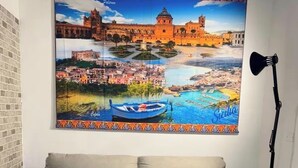 Ein beiges Sofa mit zwei Kissen, ein farbenfroher Teppich mit Muster und ein großes gerahmtes Bild von einer Küstenstadt.