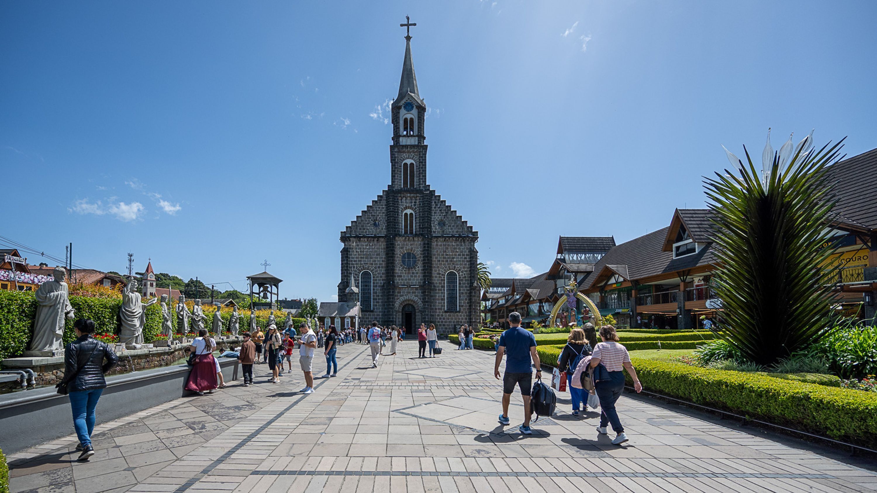A church with a tall steeple and a clock, surrounded by a plaza with people walking and statues.