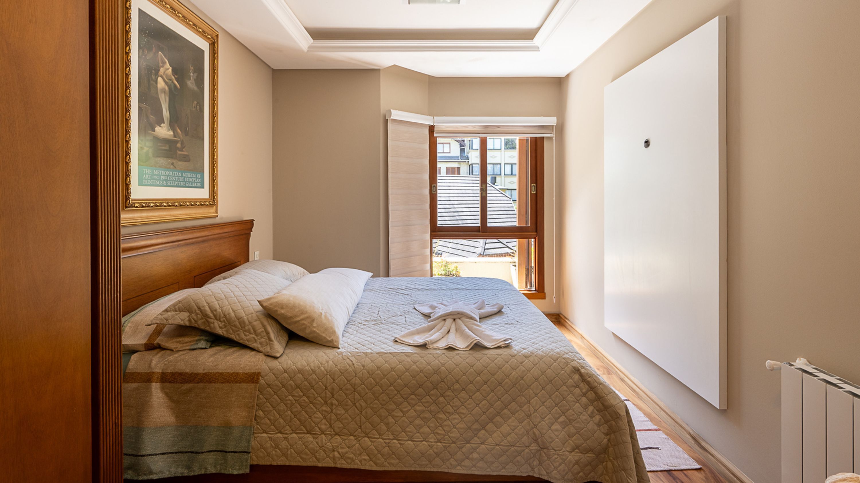 A bedroom with a bed, a painting on the wall, and a window with a view of buildings.