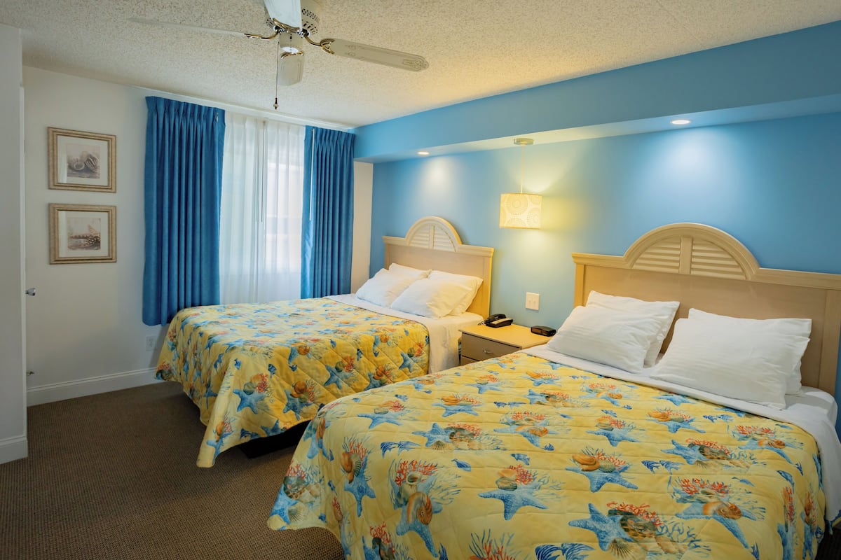 Habitación de hotel con dos camas, cada una con colcha estampada y un ventilador de techo, además de una lámpara fijada a la pared.