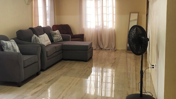 A living room with a sectional sofa, a small ottoman, a standing fan, and a flat-screen TV mounted on the wall.