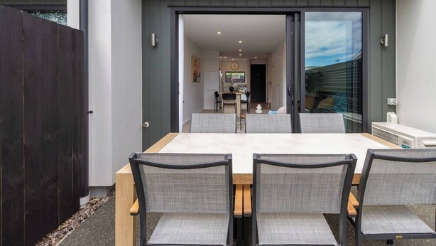 A modern outdoor dining area with a large table and four chairs, a wooden fence, and a sliding glass door leading to a kitchen. - City Stay, Sleeps 6 - Walk to One NZ Stadium (Christchurch)