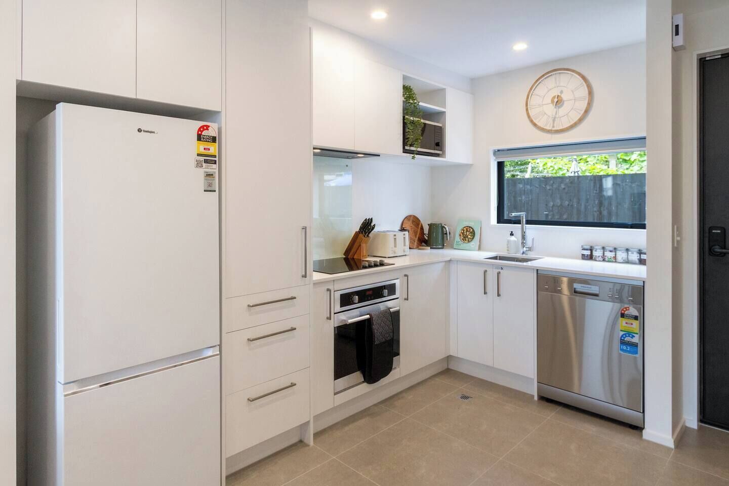 A compact kitchen with white cabinetry, a built-in oven, a dishwasher, and a window with a view of greenery.