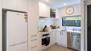 A compact kitchen with white cabinetry, a built-in oven, a dishwasher, and a window with a view of greenery. - City Stay, Sleeps 6 - Walk to One NZ Stadium (Christchurch)
