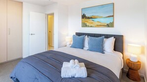 A hotel room with a large bed, a painting on the wall, and a door leading to another room. - Stadium & Port Hills View 1BR Apartment | Balcony (Christchurch)