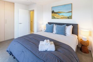 A hotel room with a large bed, a painting on the wall, and a door leading to another room. - Stadium & Port Hills View 1BR Apartment | Balcony (Christchurch)