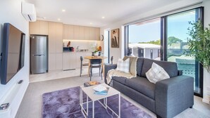 A modern living room with a grey sofa, a coffee table, and a view of the city through large windows. - Stadium & Port Hills View 1BR Apartment | Balcony (Christchurch)