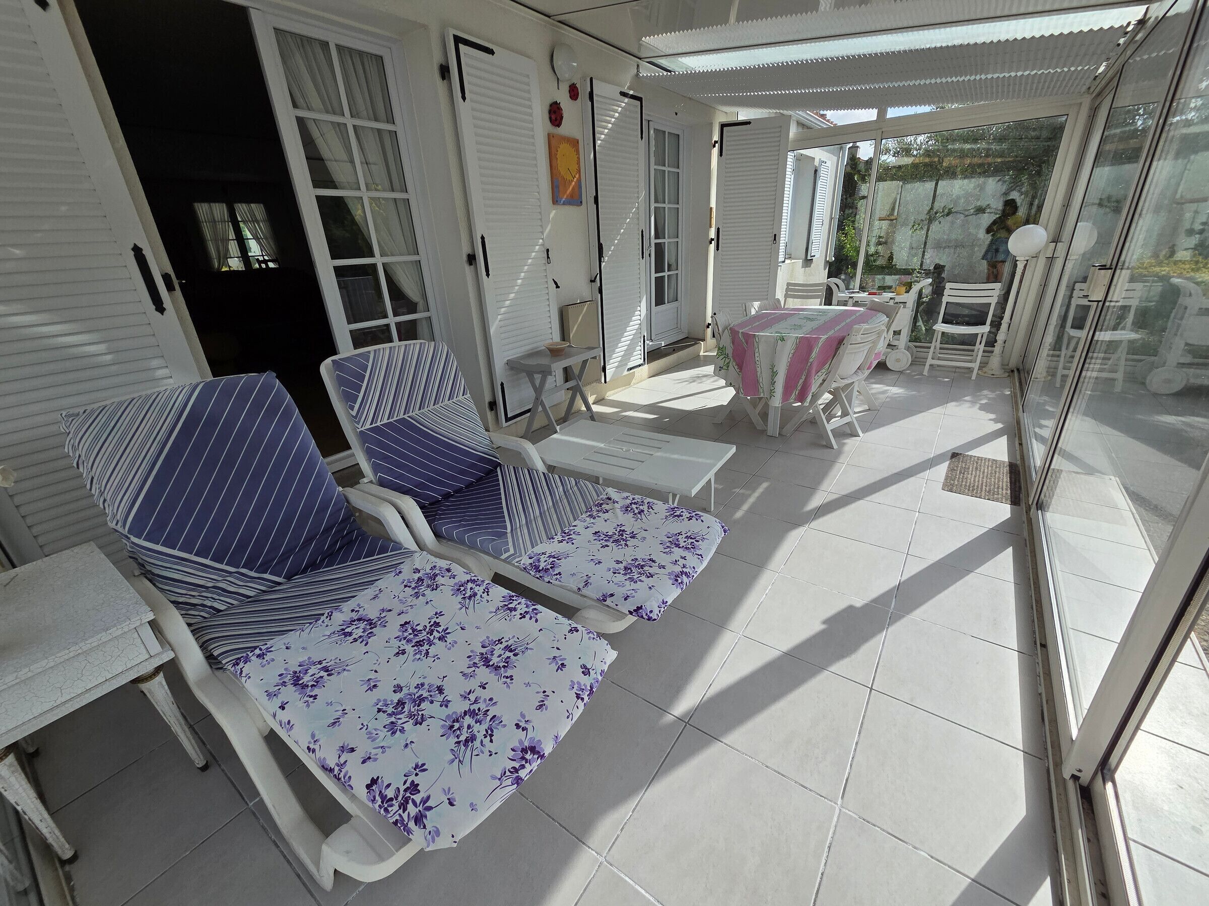 A sunlit patio with striped lounge chairs and a table, white shutters, and a glass door leading outside.