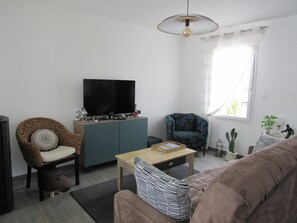A living room with a television, a sofa, a chair, a table, and a window with curtains. - Recent house near city center, 6 sleeps (La Tranche-sur-Mer)