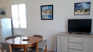 A dining area with a wooden table and chairs, a television on a cabinet, and artwork on the walls. - Single-storey cottage near beach with parking in L'Aiguillon-sur-Mer (L'Aiguillon-sur-Mer)