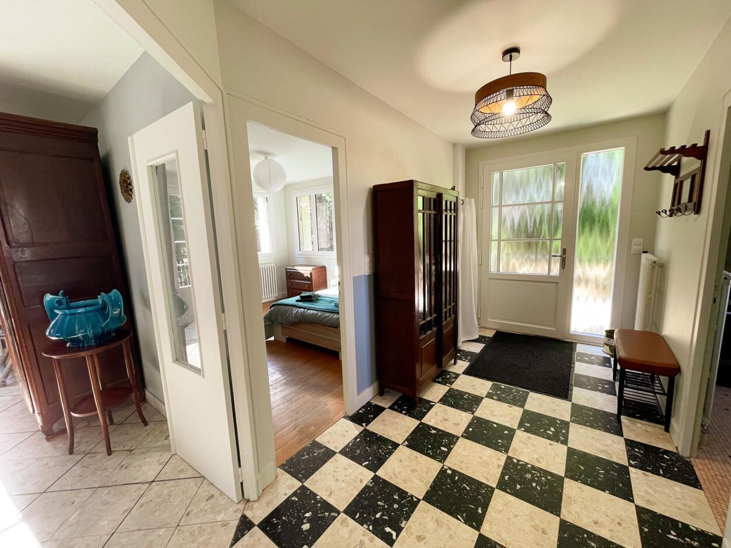 A hallway with a checkered floor, a wooden door, a blue chair, a bed with a green bedspread, and a window with curtains.