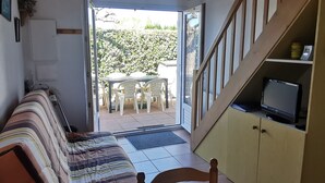 Ein Zimmer mit einer Couch, einem Fernseher auf einem Regal und Blick auf eine Terrasse mit Stühlen und einem Tisch.