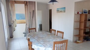 A dining area with a table covered in newspapers, wooden chairs, a bookshelf, and a view of the sea through an open door. - House for 8 near beach, pets allowed (La Tranche-sur-Mer)