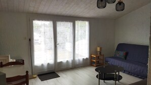 A living room with a sofa, a round table, and a chair. There are two windows with curtains, a door, and a ceiling with hanging lights. - Quiet house near beach with parking and pets allowed (La Tranche-sur-Mer)