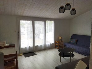 A living room with a sofa, a round table, and a chair. There are two windows with curtains, a door, and a ceiling with hanging lights. - Quiet house near beach with parking and pets allowed (La Tranche-sur-Mer)