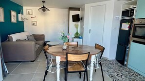 A compact living space with a dining table surrounded by chairs, a sofa, a TV mounted on the wall, and a refrigerator. - Cozy house for 4, near center and beach, with parking (La Tranche-sur-Mer)