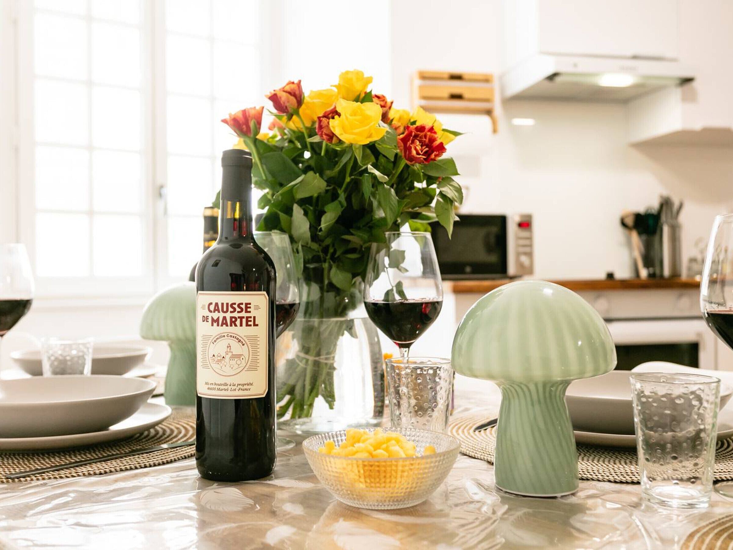 Een eettafel met een fles wijn, een bloemenvaas en een schaal fruit.