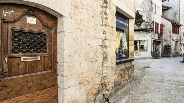 A narrow street with a wooden door marked '13' and a stone building with a shop window displaying clothing.