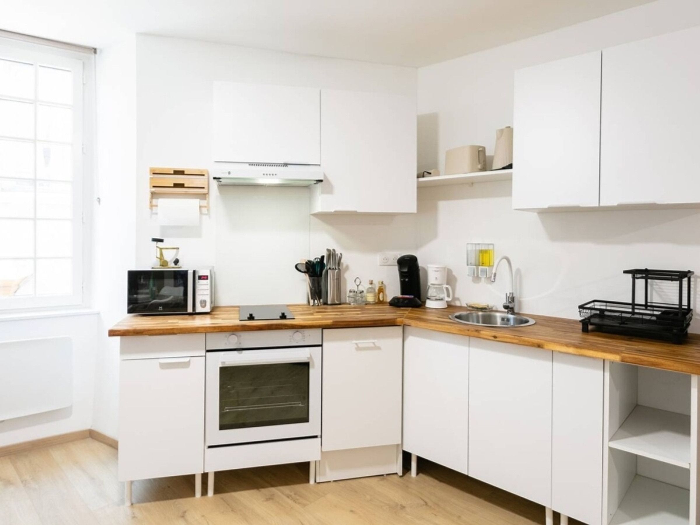 Une cuisine compacte avec des meubles blancs, un plan de travail en bois, un four encastré, un four à micro-ondes, une machine à café et un évier.