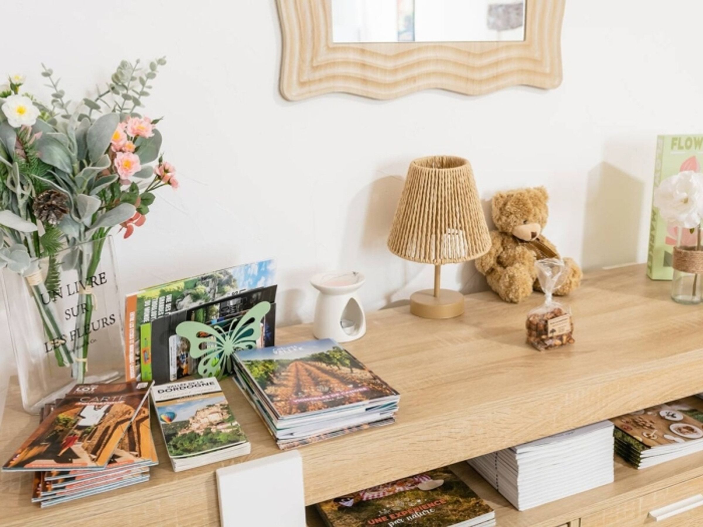 Une table en bois sur laquelle se trouve une pile de magazines, un vase avec des fleurs, une lampe, un ours en peluche et un tableau encadré accroché au mur.