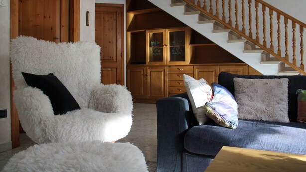 Un salón con un sillón blanco y mullido, un sofá azul con varios cojines, y una escalera de madera que lleva a una zona de cocina con armarios de madera.