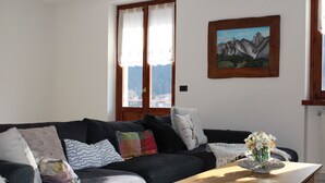 Una sala de estar con un sofá de color gris oscuro, varios cojines, una mesa de centro de madera con un jarrón de flores y un cuadro de montañas en la pared.