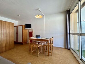 A compact living space with a dining table, chairs, a TV on a stand, and a large window providing natural light. - Studio for 4 near slopes with free wifi and parking (Le Dévoluy)