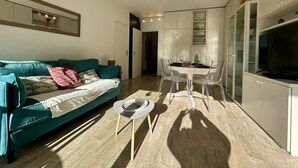 Un salon moderne avec un canapé de couleur turquoise, une table à manger entourée de chaises transparentes et un téléviseur posé sur un meuble.