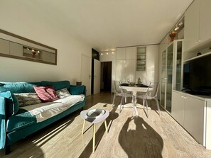 A modern living room with a teal sofa, a dining table with clear chairs, and a TV on a cabinet. - 2-Room Apt in Villeneuve: AC, Sleeps 4 (Villeneuve-Loubet)