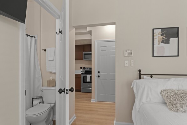 A bedroom with a bed, a fluffy pillow, a door leading to a bathroom, and a kitchen in the background.