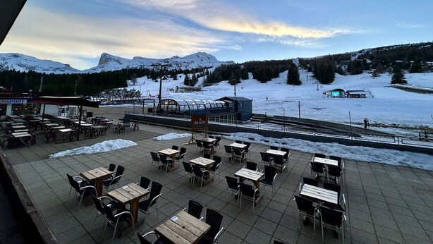 Eine Terrasse mit Tischen und Stühlen, mit Blick auf eine verschneite Landschaft mit Bergen und einer Skipiste.