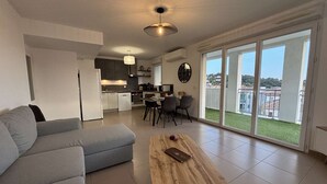 Een modern appartement met een grijze bank, een houten salontafel, een eethoek met stoelen, een keuken en grote schuifpuien naar een balkon.