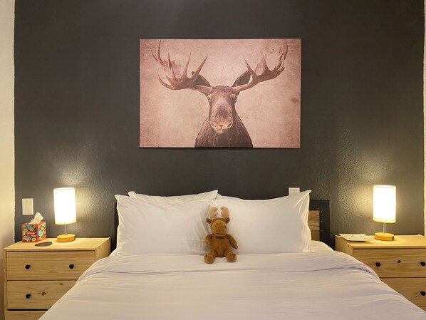 Une chambre d’hôtel avec un lit, deux tables de chevet, un ours en peluche sur le lit et un tableau représentant un cerf accroché au mur.