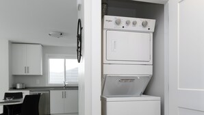 A compact kitchen with a built-in washer and dryer, a dining area with a table and chairs, and a window providing natural light. - Cozy Corner on Trumpeter (Colwood)