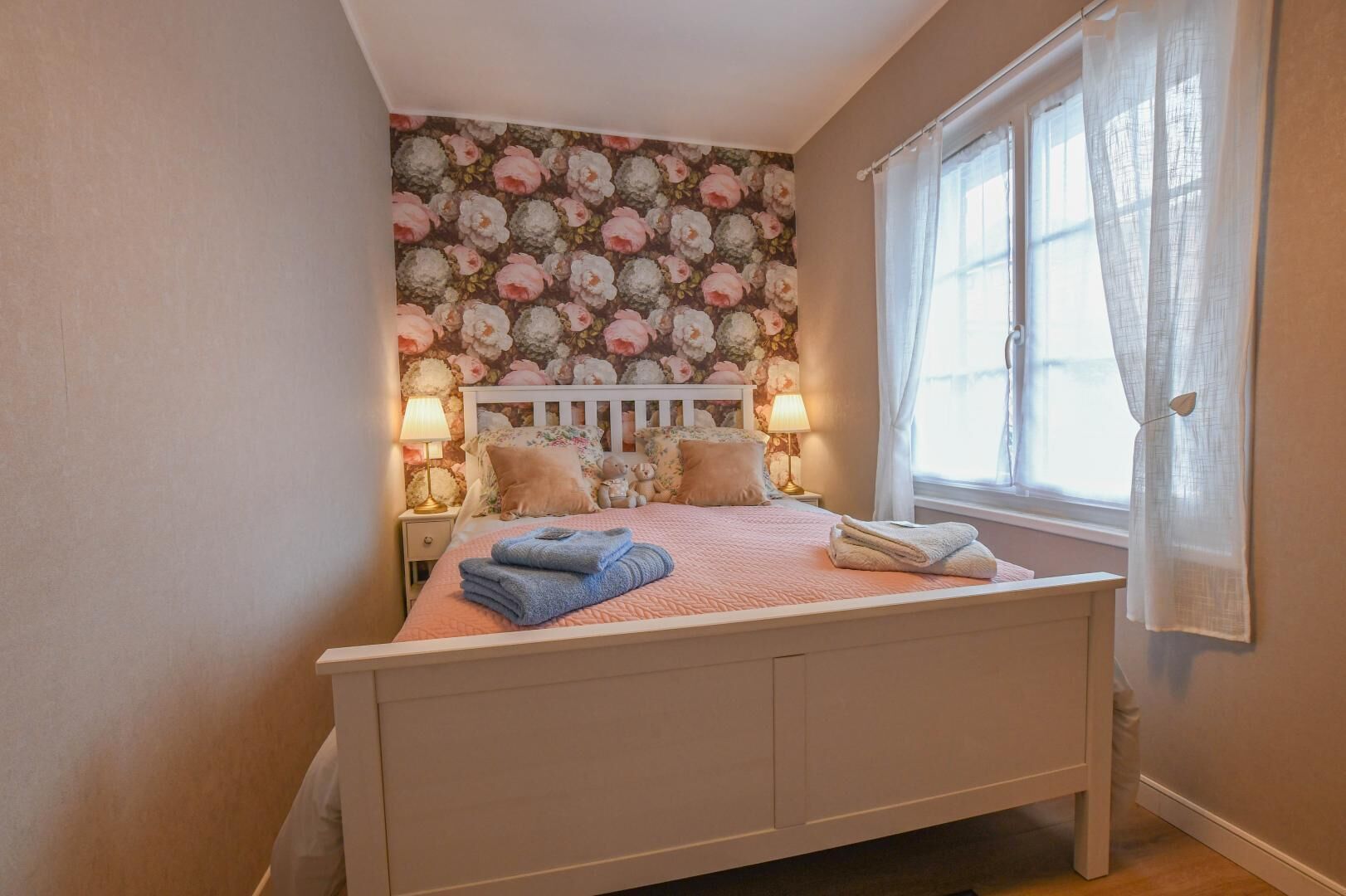 A bedroom with a floral wallpaper, a white bed frame, two bedside lamps, and a window with sheer curtains.