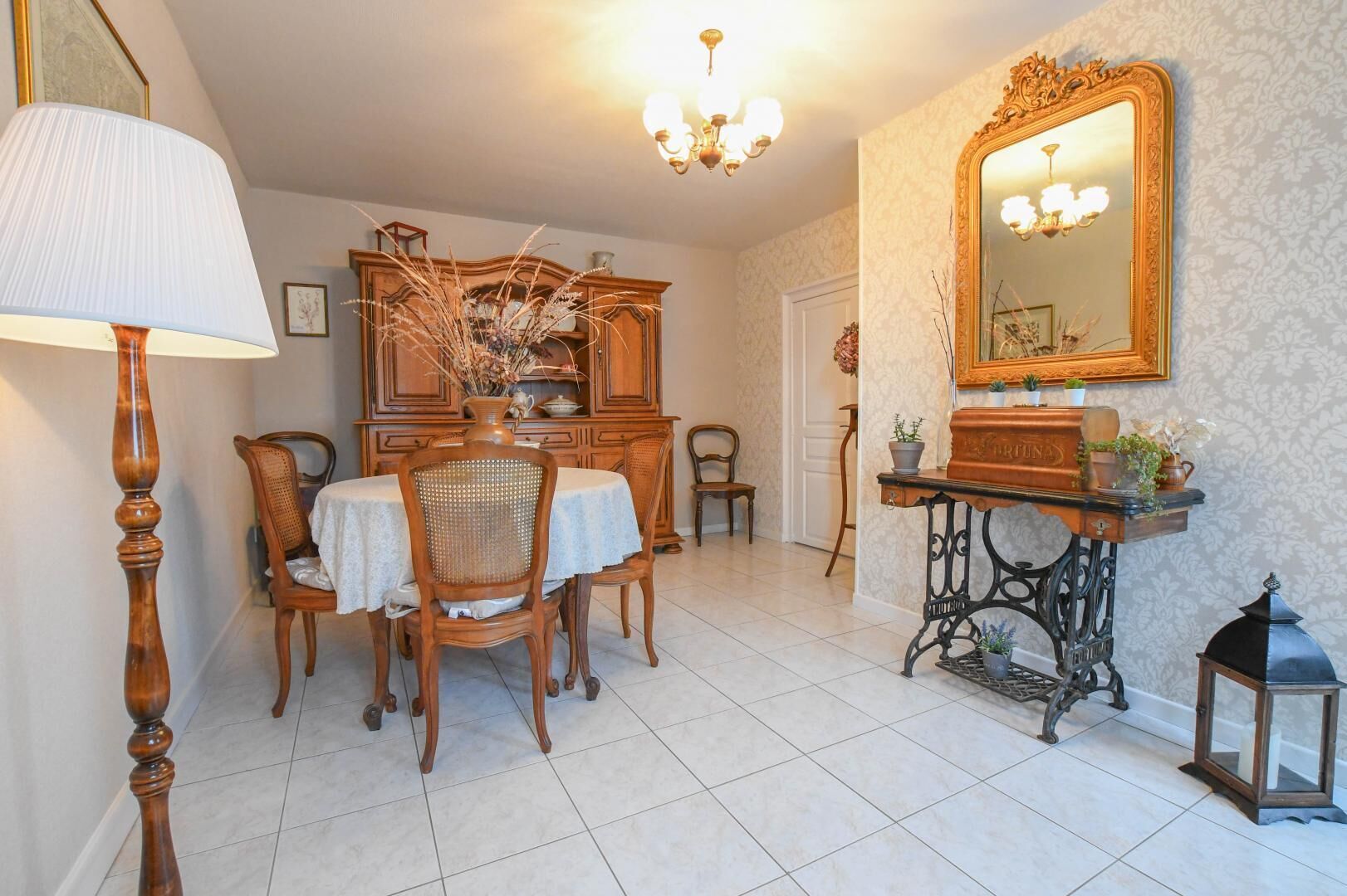 A dining area with a wooden table and chairs, a vase with dried plants, a chandelier, a mirror, and a vintage sewing machine.