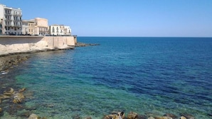 Un edificio costero de varios pisos, con vista despejada al mar y terreno rocoso en la orilla.