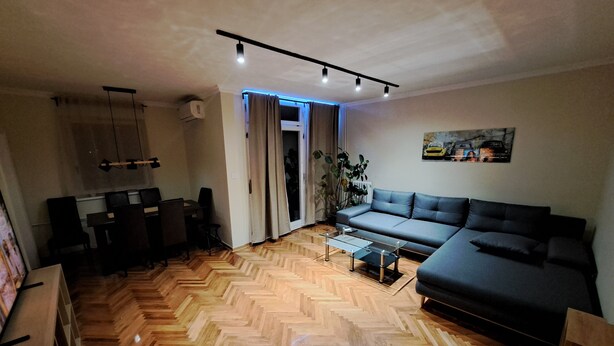 A living room with a herringbone wooden floor, a grey sofa, a glass coffee table, and a dining area with chairs and a table. - Cozy apartment in lovely and peaceful part of Belgrade (Belgrade)