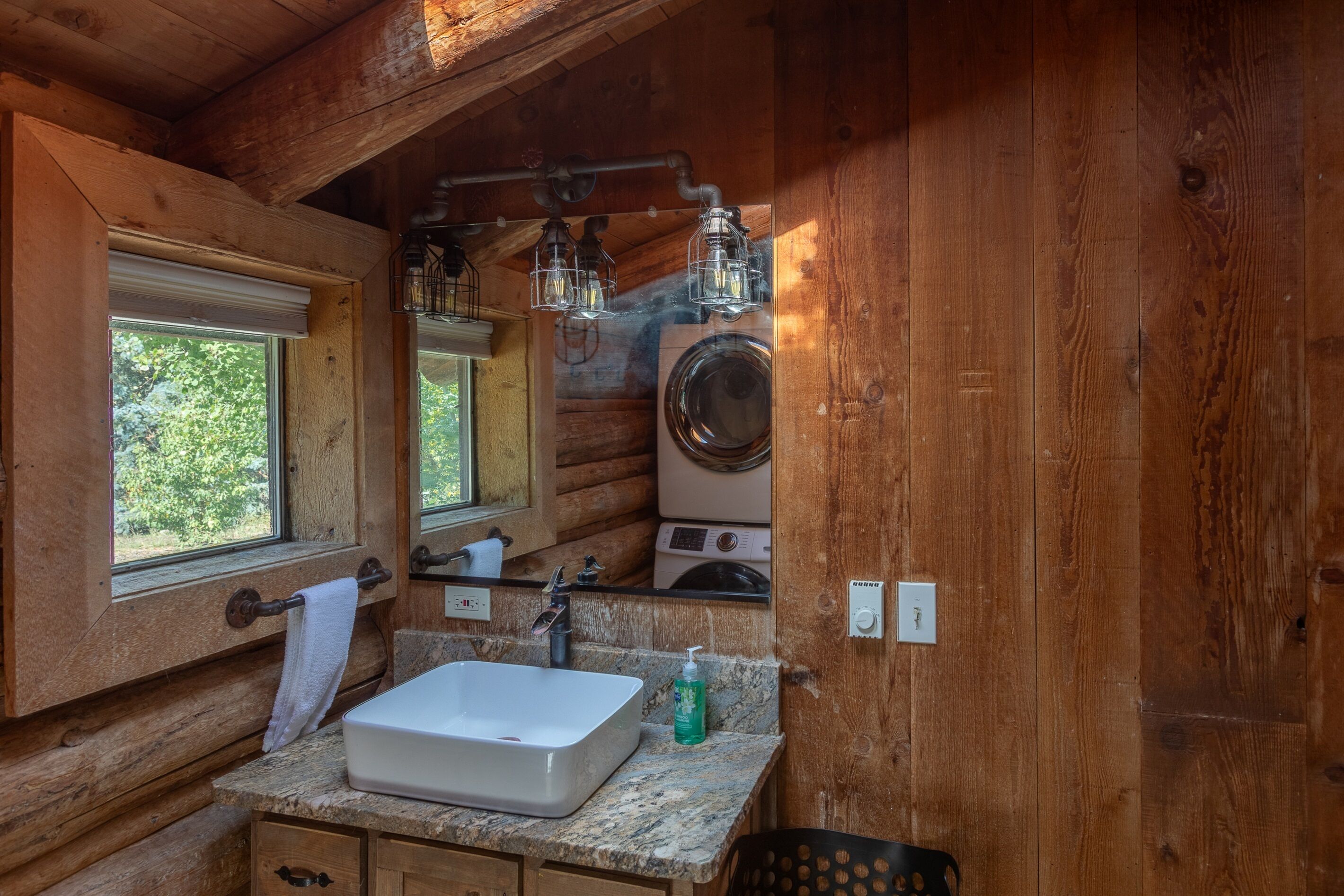 A wooden cabin bathroom with a sink, mirror, towel rack, and a washing machine.