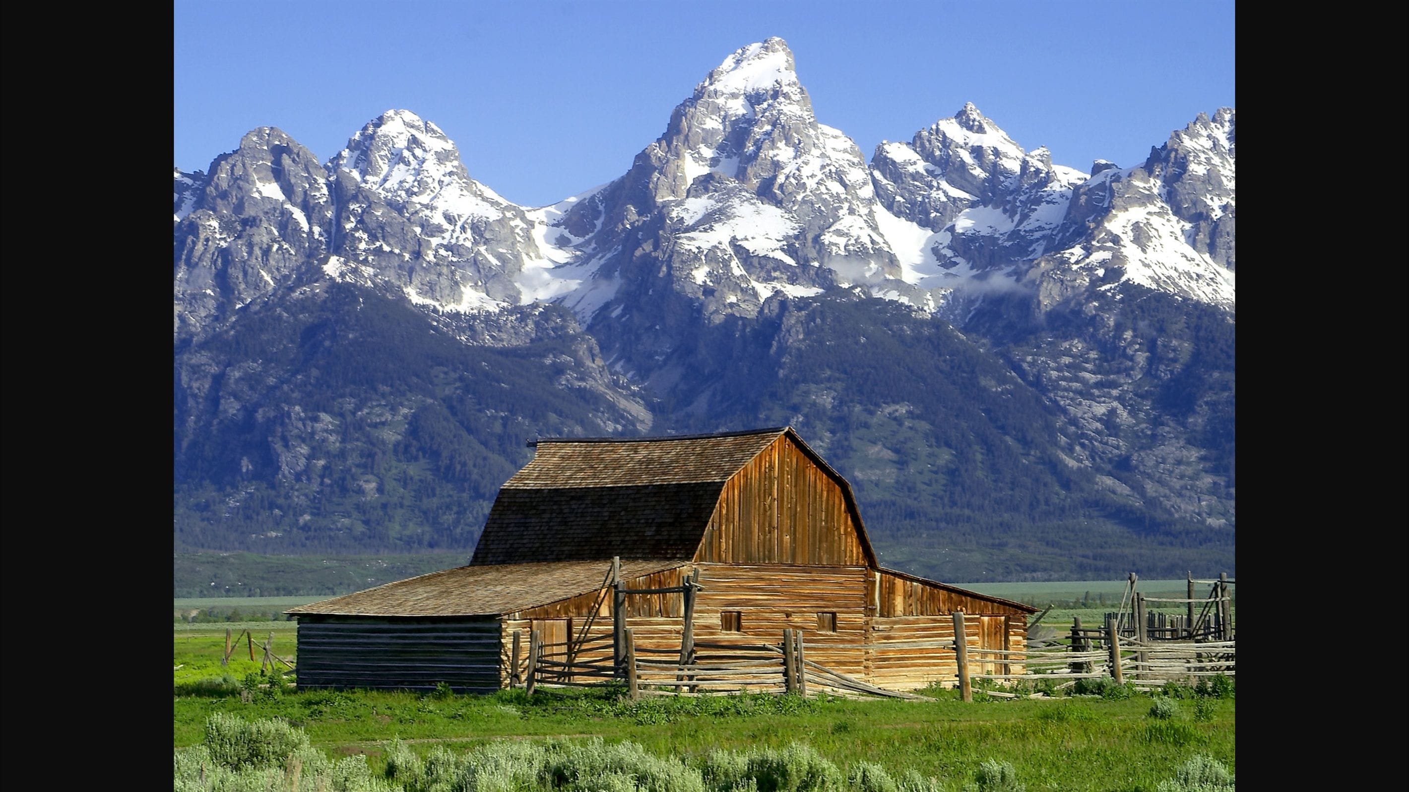 A wooden barn with a sloped roof, surrounded by a wooden fence, set against a backdrop of snow-capped mountains.