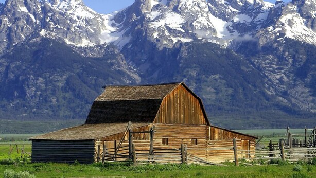 Une grange en bois, dotée d’un toit en pente et entourée d’une clôture en bois, se dressant sur fond de montagnes enneigées.