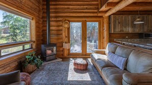 L’intérieur d’une cabane en rondins, avec une cheminée, un canapé et de grandes fenêtres donnant sur le paysage extérieur.