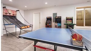 Ein Tischtennistisch mit Netz, ein Basketballkorb und Spielautomaten in einem Raum mit Tür und Fenstern.