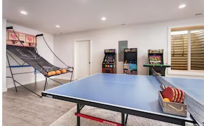 A ping pong table with a net, a basketball hoop, and arcade games in a room with a door and windows.
