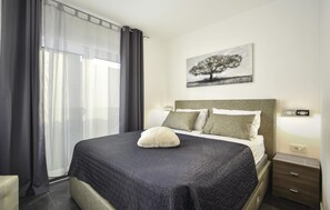 Un lit bien fait, avec une couverture sombre, un oreiller blanc moelleux et deux oreillers recouverts d’un tissu texturé. Un tableau représentant un arbre est accroché au mur, au-dessus du lit.