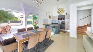 Una cocina moderna con una isla central, un comedor con una mesa y sillas de madera, y una escalera que sube a un nivel superior.