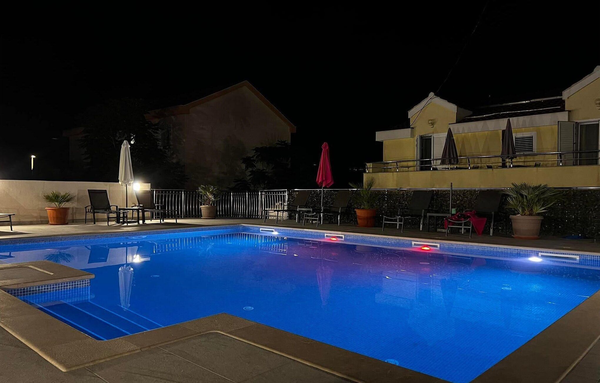 Una piscina con tumbonas y sombrillas, rodeada de plantas en macetas y un edificio con balcones.