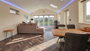 A spacious living room with a herringbone floor, a sofa, a dining table, and a view of a garden through large sliding glass doors. - Hunters Cottage – A Romantic Luxury Retreat with Hot Tub & Slipper Bath, AC (Blandford Forum)