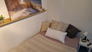 A bedroom with a bed, pillows, a nightstand, a lamp, a remote control, and a wall-mounted picture of a beach scene. - Cozy 1-room apartment in the Hildesheim forest (Diekholzen)